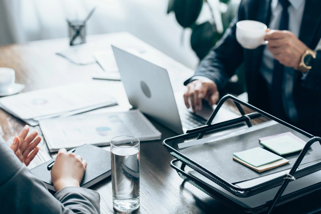 Cropped view of businesswoman holding pen near notebook, glass of water and investor with cup of coffee in office