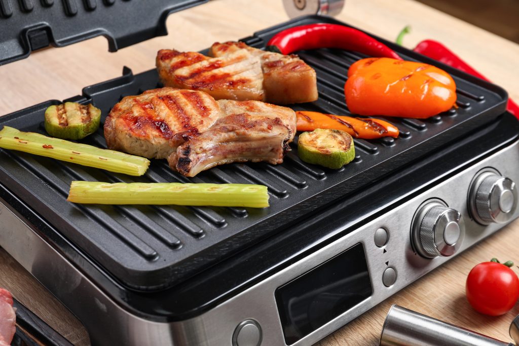 Modern electric grill with tasty steaks and vegetables on wooden background, closeup