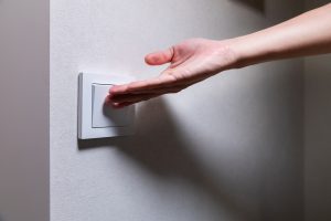 Close-up of an unrecognizable young Caucasian womans hand turning on a light in a dark room by pressing a switch on a white wall. Close-up.